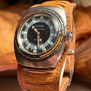 Puede incluir: Un reloj de pulsera plateado con una esfera negra y detalles en naranja. El reloj tiene una correa de cuero marr&oacute;n con un dise&ntilde;o cosido. La esfera del reloj dice "Восток  магнитные  18 кал  Сделано в СССР".