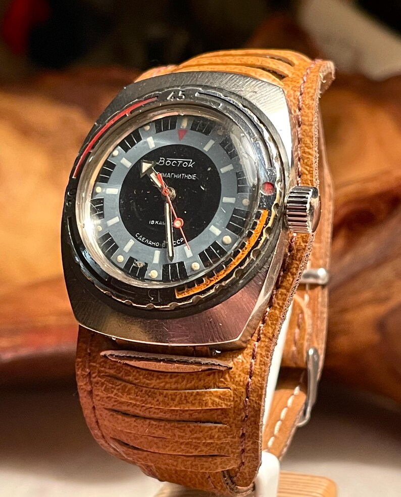 Puede incluir: Un reloj de pulsera plateado con una esfera negra y detalles en naranja. El reloj tiene una correa de cuero marr&oacute;n con un dise&ntilde;o cosido. La esfera del reloj dice "Восток  магнитные  18 кал  Сделано в СССР".
