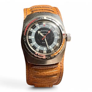 Puede incluir: Un reloj de pulsera plateado con una esfera negra y una correa de cuero marr&oacute;n. La esfera del reloj tiene marcas blancas y el texto "Восток АВТОМАТИЧЕСКИЕ" en escritura cir&iacute;lica.