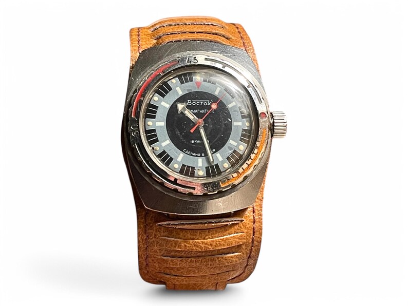 Puede incluir: Un reloj de pulsera plateado con una esfera negra y una correa de cuero marr&oacute;n. La esfera del reloj tiene marcas blancas y el texto "Восток АВТОМАТИЧЕСКИЕ" en escritura cir&iacute;lica.