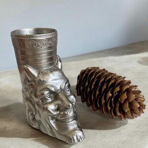 May include: A silver metal goblet shaped like a devil's head with a textured surface. The goblet is sitting next to a brown pine cone.