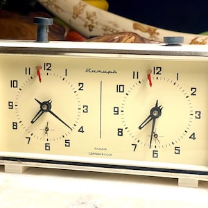 May include: A rectangular white analog chess clock with two circular clock faces featuring black numerals and hands. The device has two blue top-mounted buttons, a silver-toned frame, and Cyrillic text on the face reading Yantar, 4 kamnya, Sdelano v SSSR.