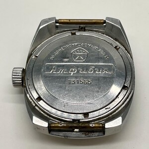 Puede incluir: La parte posterior de un reloj de pulsera plateado con el texto "ВОДОНЕПРОНИЦАЕМЫЕ 200 М" y "Амфибия" impreso en la caja. El reloj tiene el n&uacute;mero de serie "31555".