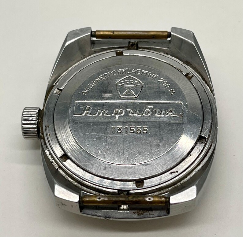 Puede incluir: La parte posterior de un reloj de pulsera plateado con el texto "ВОДОНЕПРОНИЦАЕМЫЕ 200 М" y "Амфибия" impreso en la caja. El reloj tiene el n&uacute;mero de serie "31555".