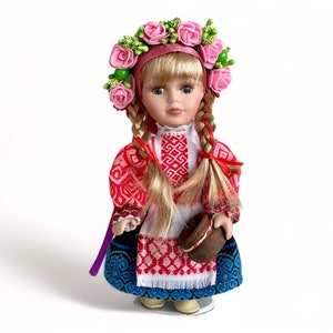 May include: A porcelain doll wearing a traditional Ukrainian folk costume with a red and white embroidered blouse, a blue and white skirt, and a pink flower crown. The doll is holding a small brown basket.