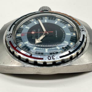 Puede incluir: Un reloj de pulsera plateado con una esfera negra y un bisel rojo y blanco. El reloj tiene una ventana de fecha y el texto "BOСTOK" y "СДЕЛАНО В СССР" en la esfera.