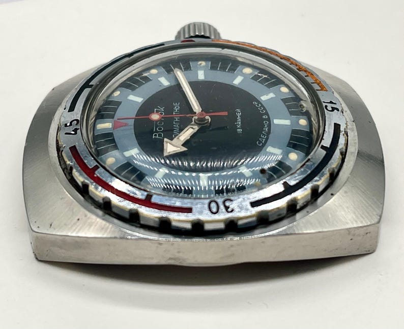 Puede incluir: Un reloj de pulsera plateado con una esfera negra y un bisel rojo y blanco. El reloj tiene una ventana de fecha y el texto "BOСTOK" y "СДЕЛАНО В СССР" en la esfera.