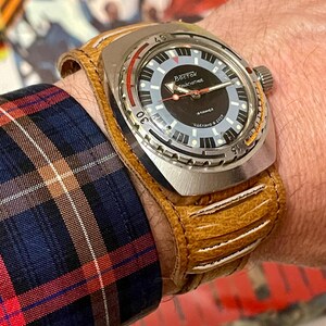 Puede incluir: Un reloj de pulsera plateado con una esfera negra y una correa de cuero marr&oacute;n. La esfera del reloj tiene marcas blancas y el texto "Восток ЧАСОВЫЕ" en escritura cir&iacute;lica.