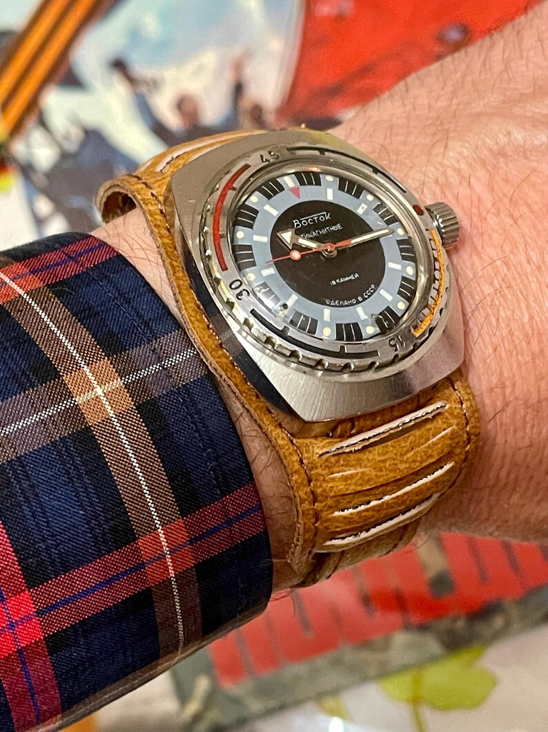 Puede incluir: Un reloj de pulsera plateado con una esfera negra y una correa de cuero marr&oacute;n. La esfera del reloj tiene marcas blancas y el texto "Восток ЧАСОВЫЕ" en escritura cir&iacute;lica.