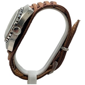Puede incluir: Un reloj de pulsera plateado con una correa de cuero marr&oacute;n. El reloj tiene una esfera redonda con un bisel negro y una corona plateada.