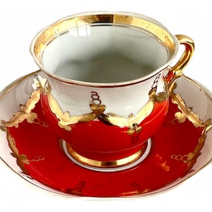 May include: A vintage teacup and saucer set. The cup and saucer are white with red and gold accents. The cup has a gold handle and rim. The saucer has a red center and gold trim. The set is likely porcelain.