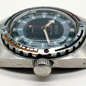 Puede incluir: Un reloj de pulsera plateado con una esfera negra y una aguja de segundos roja. El reloj tiene un bisel giratorio con marcas negras y blancas y la palabra "BOCTAK" impresa en la esfera.