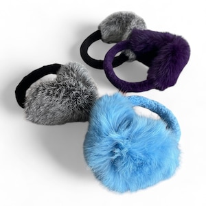 May include: Three pairs of faux fur earmuffs in gray, purple, and light blue. The earmuffs are made of soft, fluffy fur and have a headband that goes around the head.