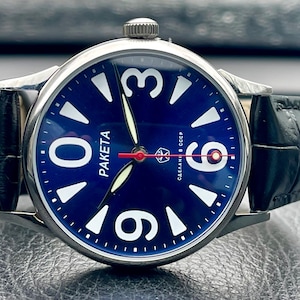 Puede incluir: Un reloj de pulsera vintage con esfera azul oscuro, grandes números blancos y la palabra "PAKETA" en blanco. El reloj tiene una caja plateada, una correa de cuero negro y un segundero rojo. También es visible el texto "СДЕЛАНО В СССР".