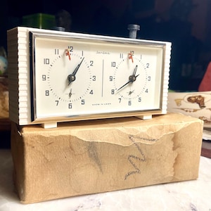 May include: A rectangular analog chess clock with a white casing and two circular clock faces featuring black numerals and hands. The clock has two black top buttons, a silver-toned frame, and the text Jantar and MADE IN USSR on the face.