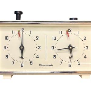 May include: A vintage chess clock with a cream face and a silver frame. It features two dials with black numerals and hands, and a red second hand. The clock has a black button on top and the word "Ястемь" on the bottom.