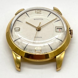 May include: A gold-tone wristwatch with a white face and black hands. The watch face has the word "Wostok" and "18 jewels" printed on it.