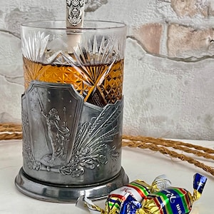 May include: A silver tea cozy with an ornate design, holding a glass of tea with a silver spoon. Two wrapped candies are on a white surface.