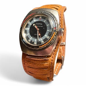 May include: A silver-toned wristwatch with a black face and a brown leather band. The watch face has white markings and the text "BOCTON AВTOMATИЧEСКИЕ" in Cyrillic script.
