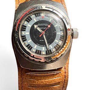 Puede incluir: Un reloj de pulsera vintage plateado con una esfera negra y detalles rojos. El reloj tiene una correa de cuero marr&oacute;n con un patr&oacute;n texturizado. La esfera del reloj tiene el texto "Восток АМФИБИЯ 18 КАЛ СДЕЛАНО В СССР" impreso en &eacute;l.