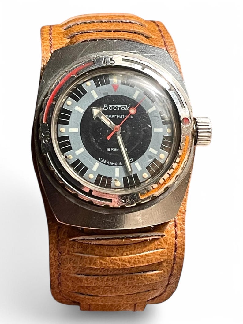 Puede incluir: Un reloj de pulsera vintage plateado con una esfera negra y detalles rojos. El reloj tiene una correa de cuero marr&oacute;n con un patr&oacute;n texturizado. La esfera del reloj tiene el texto "Восток АМФИБИЯ 18 КАЛ СДЕЛАНО В СССР" impreso en &eacute;l.