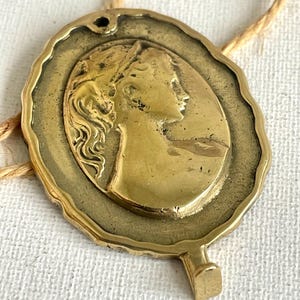 Peut inclure: Pendentif en laiton de style vintage avec le profil d'une femme en relief. Le pendentif ovale a un bord texturé et un petit trou en haut pour la suspension. Un petit crochet est en bas. Il est attaché à une ficelle naturelle.