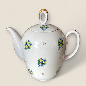Puede incluir: Tetera de porcelana blanca con tapa y asa doradas. La tetera está decorada con diseños florales azules y amarillos.