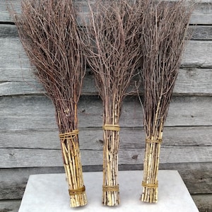 May include: Three brown twig brooms with natural twine bindings. The brooms are tied together at the top and are standing upright on a white surface.