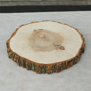 May include: A round wooden slice with a natural bark edge. The wood is light brown and has a natural grain pattern. The slice is perfect for crafts, decor, or as a serving tray.