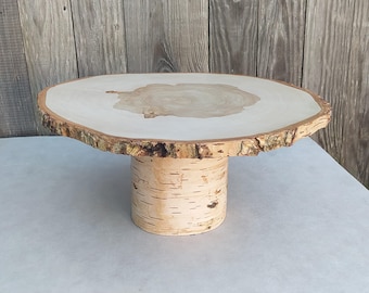 May include: A natural wood cake stand with a round top and a cylindrical base. The top is a light color with a darker center, and the edge has a rough, bark-like texture. The base is made of birch bark.