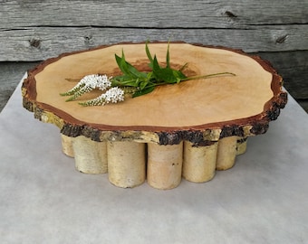 May include: A wooden cake stand with a natural bark edge and a birch bark base. The stand is decorated with white flowers.