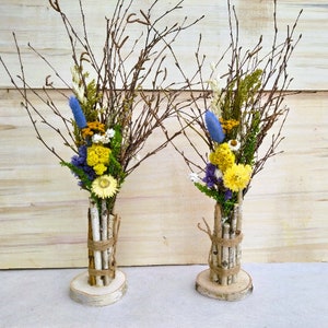 May include: Two decorative floral arrangements. Each features a bundle of dried branches and colourful dried flowers, including yellow, blue, and white blooms. The base is a small wooden slice, and the stems are wrapped with birch bark and twine.
