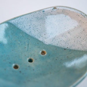 Könnte beinhalten: Handgefertigte Seifenschale aus Keramik in Tropfenform. Die Schale hat eine gesprenkelte türkisfarbene Farbe mit einem weißen, gesprenkelten Abschnitt oben. Drei Abflusslöcher sind sichtbar.