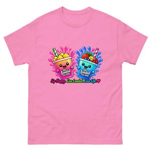 Puede incluir: Una camiseta rosa con un gráfico de dos tazas de dibujos animados con caras, una naranja y otra azul, rodeadas de frutas y salpicaduras de líquido. El texto dice "the loaded tea shop" y "Sip Happy. Live Loaded. Love Life." en la parte inferior.