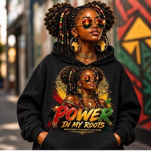 Op de afbeelding: Zwarte hoodie met een levendige afbeelding van een vrouw met gevlochten haar, zonnebril en gouden sieraden. De afbeelding bevat de tekst "POWER IN MY ROOTS" in rood en groen, met extra tekst eronder. De vrouw draagt een zonnebril met de kleuren van de Rastafari-vlag.