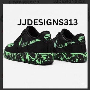 May include: Pair of black Nike Air Force 1 sneakers with neon green accents. The Nike swoosh and heel logo are in neon green. The soles and side panels feature a splatter design in neon green. The text "JJDESIGNS313" is at the top.