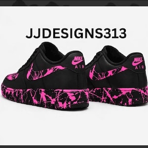 May include: Black Nike Air Force 1 sneakers with pink splatter design on the soles, swoosh, and accents. The text "JJDESIGNS313" is at the top of the image. The Nike logo is visible on the heel.
