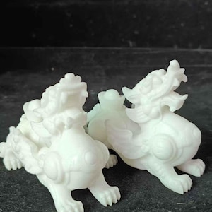 May include: Two white, translucent, carved stone figurines depicting mythical creatures with open mouths, detailed manes, and wings. Each creature is positioned in a crouching stance, showcasing intricate textures and smooth, polished surfaces.
