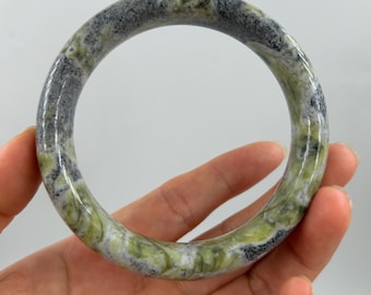 Puede incluir: Un brazalete redondo y pulido con un patrón veteado en verde, gris y blanco. El brazalete se sostiene en una mano sobre un fondo blanco. El brazalete tiene una forma circular sólida.