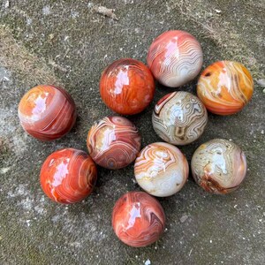 Può includere: Una collezione di dieci sfere di agata lucidata nei toni del rosso, arancione e crema. Le sfere hanno una finitura lucida e mostrano intricati motivi a bande. Sono disposte su una superficie strutturata e muschiosa.