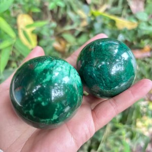 May include: Two polished, green gemstone spheres with swirling patterns are held in a hand. The spheres have a glossy finish and varying shades of green. The background is blurred, with green foliage.