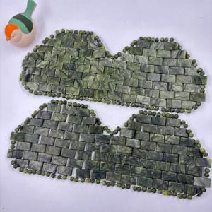Op de afbeelding: Twee jade stenen massagematten met een baksteenachtig patroon, verbonden door kleine jade kralen. De matten hebben een gevlekte groene kleur. Een kleine keramische vogel met oranje, groene en witte accenten staat in de linkerbovenhoek.