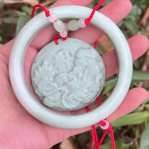 May include: A pale green jade pendant and bracelet set. The pendant features a detailed dragon carving. The bracelet is a smooth, circular bangle. Both pieces are connected by a red cord with small beads. The set is held in a hand.