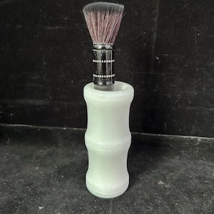 May include: A shaving brush with a light green, bamboo-style handle. The brush has a black metal collar with a decorative pattern and a dark brown bristle head. The brush is designed to stand upright.