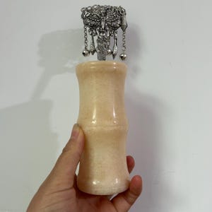May include: A decorative bottle stopper with a silver filigree design and dangling charms. The stopper is attached to a light beige, bamboo-shaped bottle. The bottle is held by a hand.