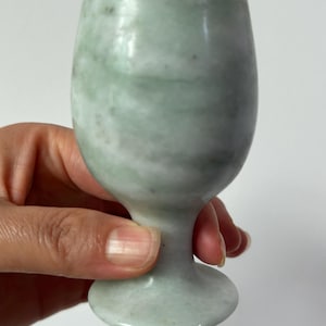 Könnte beinhalten: Ein kleiner, hellgrüner Kelch aus Stein. Der Kelch hat eine abgerundete Schale, einen schlanken Stiel und einen kreisförmigen Fuß. Der Stein hat ein marmoriertes Aussehen mit subtilen Farbvariationen. Der Kelch wird von einer Hand vor weißem Hintergrund gehalten.