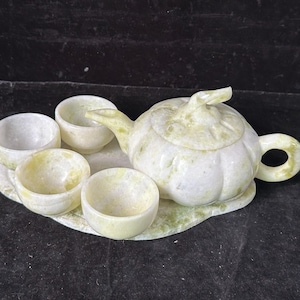 Puede incluir: Un juego de té de piedra verde claro y blanco. El juego incluye una tetera con tapa, cuatro tazas de té pequeñas y una bandeja. La tetera tiene un asa y un pico curvos. Las tazas son redondas y tienen un acabado liso.