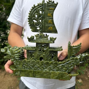May include: A carved jade boat sculpture in shades of green. The boat features intricate details, including a dragon head at the bow, a pagoda-style structure, and a sail with Chinese characters. The sculpture is held by a person.