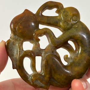 May include: A carved jade pendant featuring a monkey clutching a peach. The pendant is a mix of brown and green hues, with intricate details. The monkey is in a playful pose, and the peach is large and detailed.
