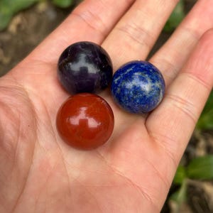 May include: Three polished gemstone spheres: a deep purple amethyst, a vibrant blue lapis lazuli, and a rich red jasper. The spheres are held in a hand, showcasing their smooth, reflective surfaces and natural colors. These decorative spheres are ideal for meditation or display.